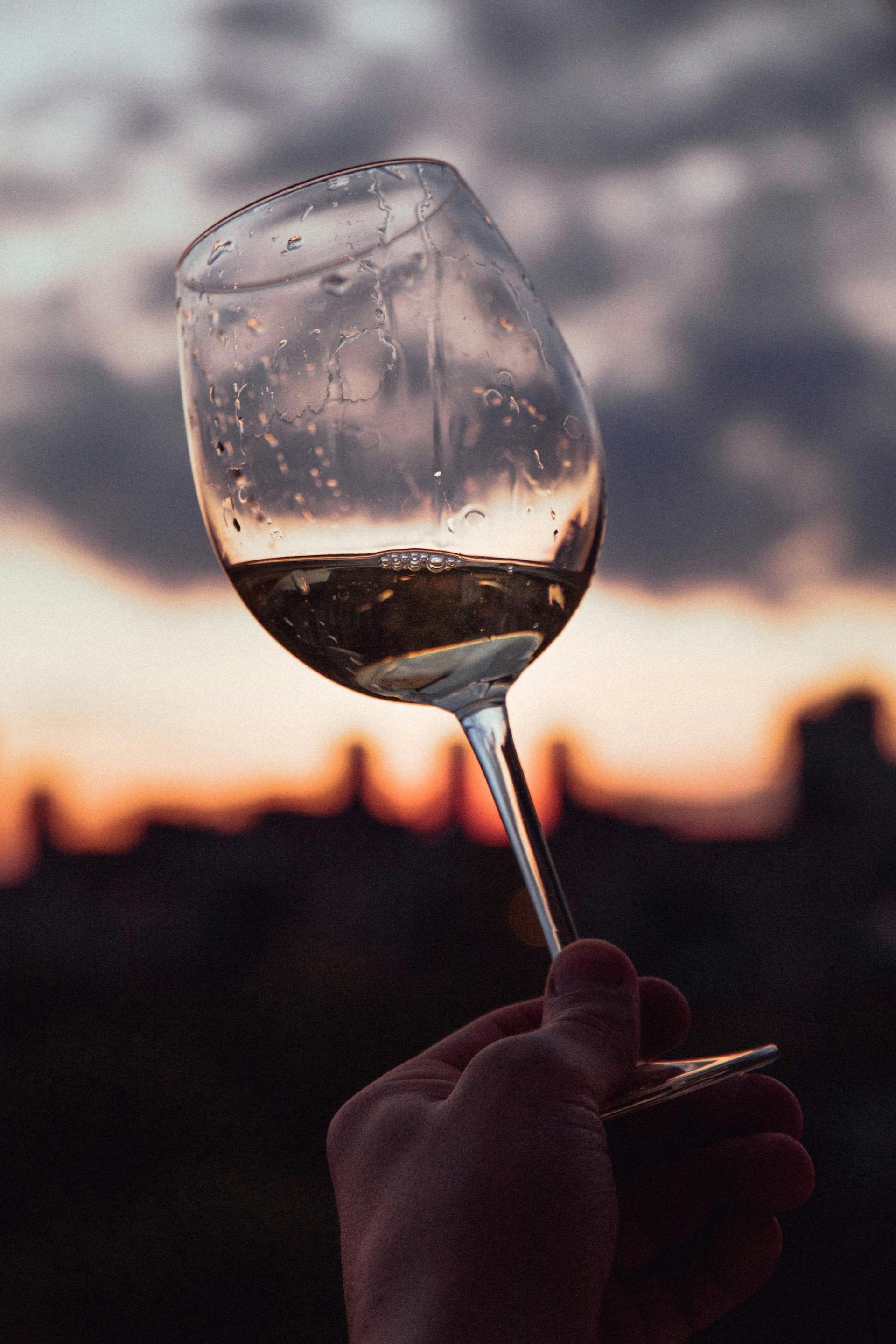 Person hält Weinglas mit klarer Flüssigkeit und Hintergrund Sonnenuntergang