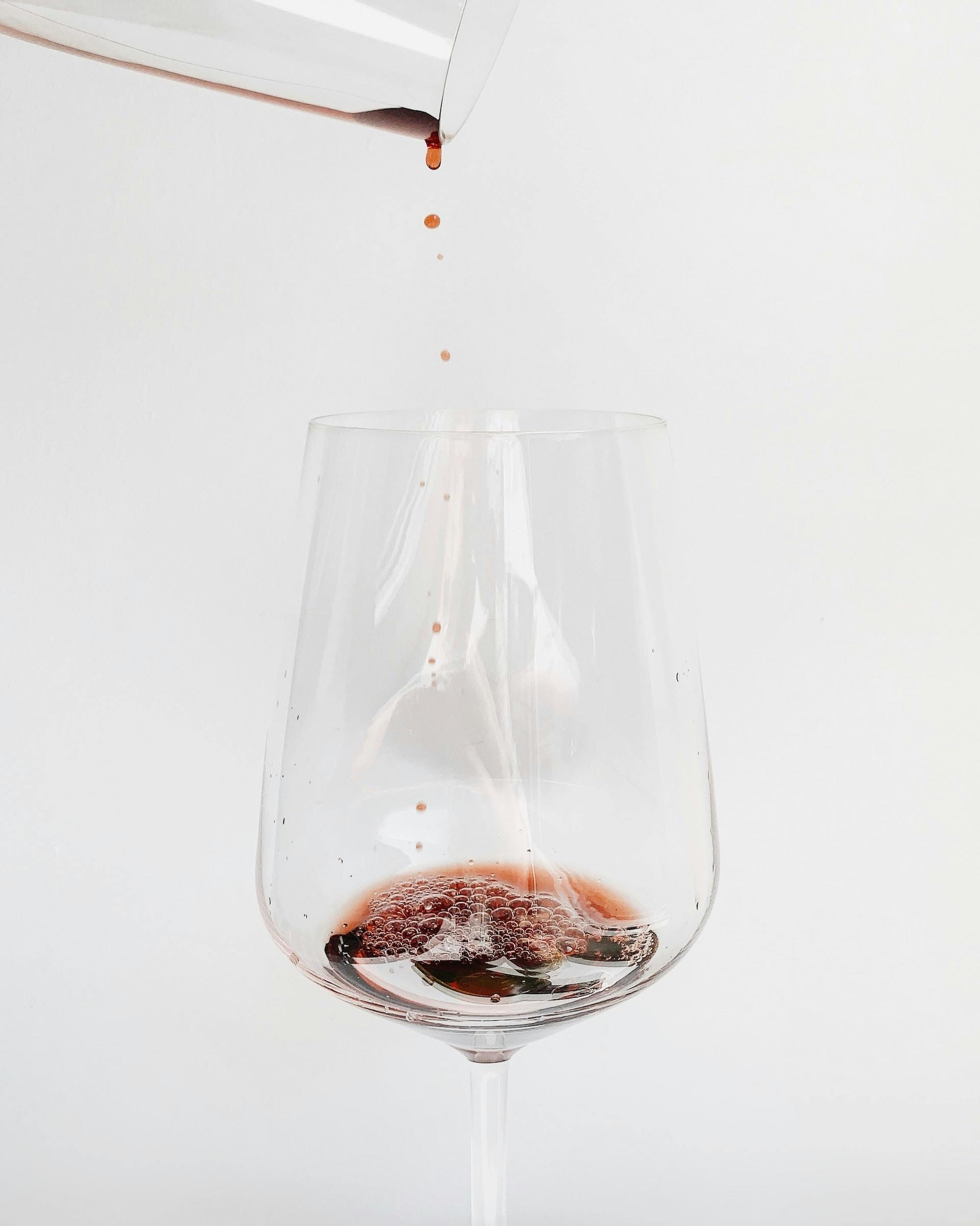 Weinglas mit Weißem hintergrund wo aus Gefäß rote Flüssigkeit reintropft