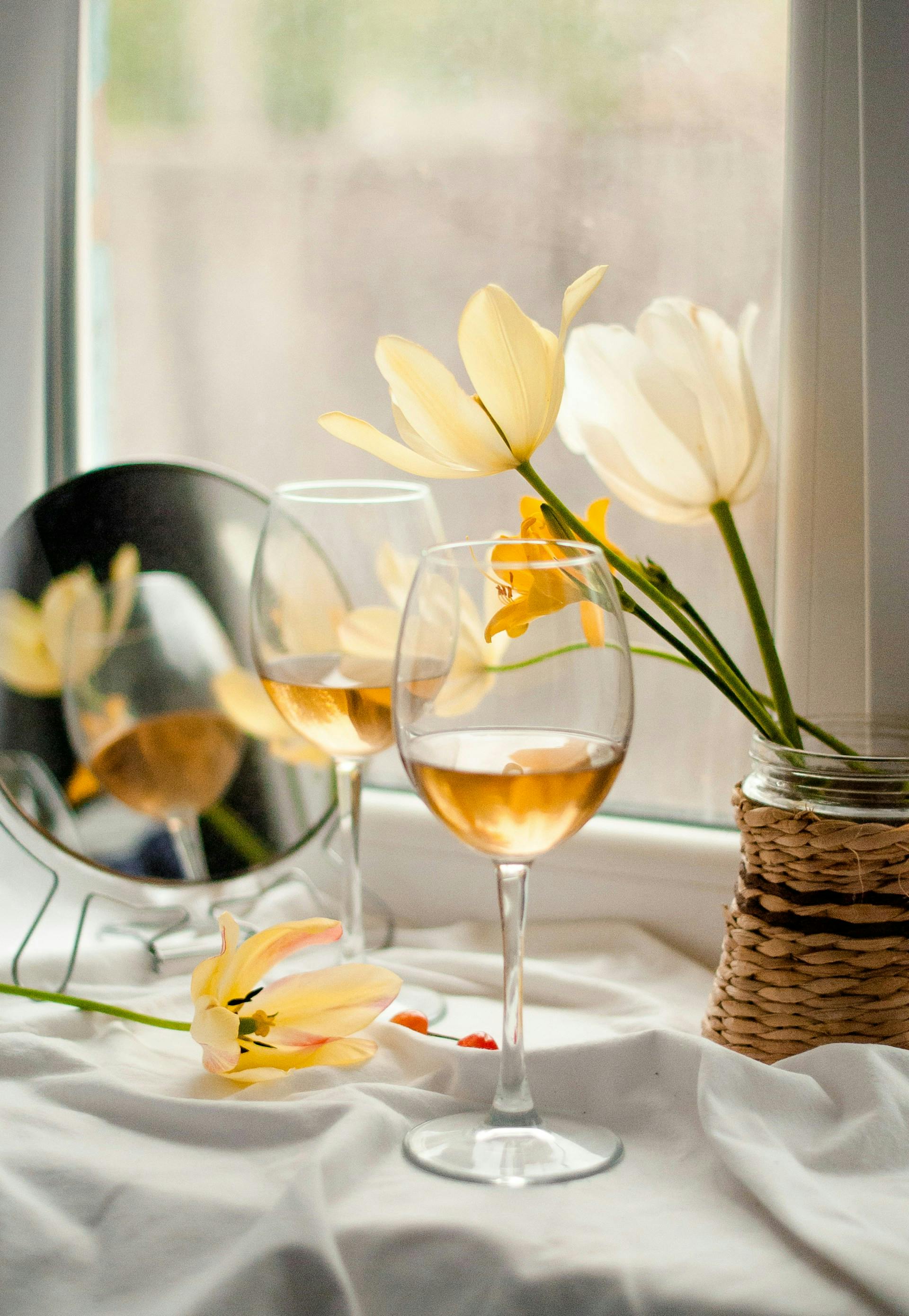 Zwei Gläser mit goldener Flüssigkeit stehen auf weißer Tischdecke neben gelben Blumen.