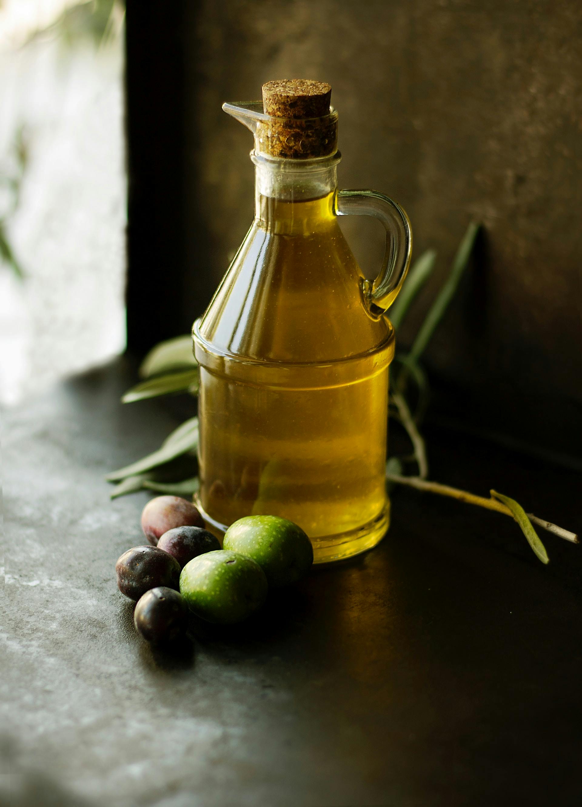 Klare Flasche mit Olivenöl und Oliven auf dem dunklen Hintergrund