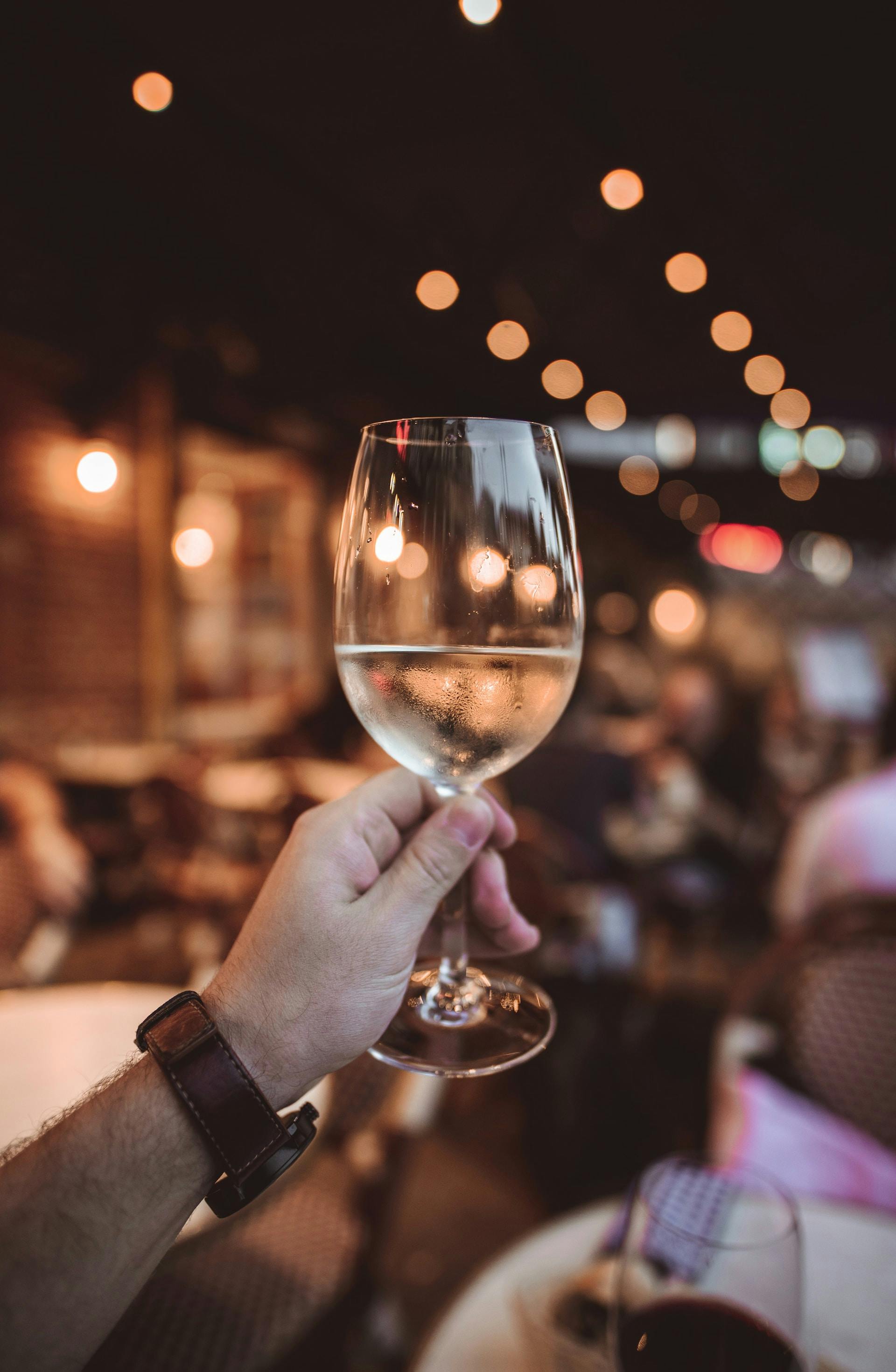 Hand hält Glas mit Weißwein, Lichter im Hintergrund