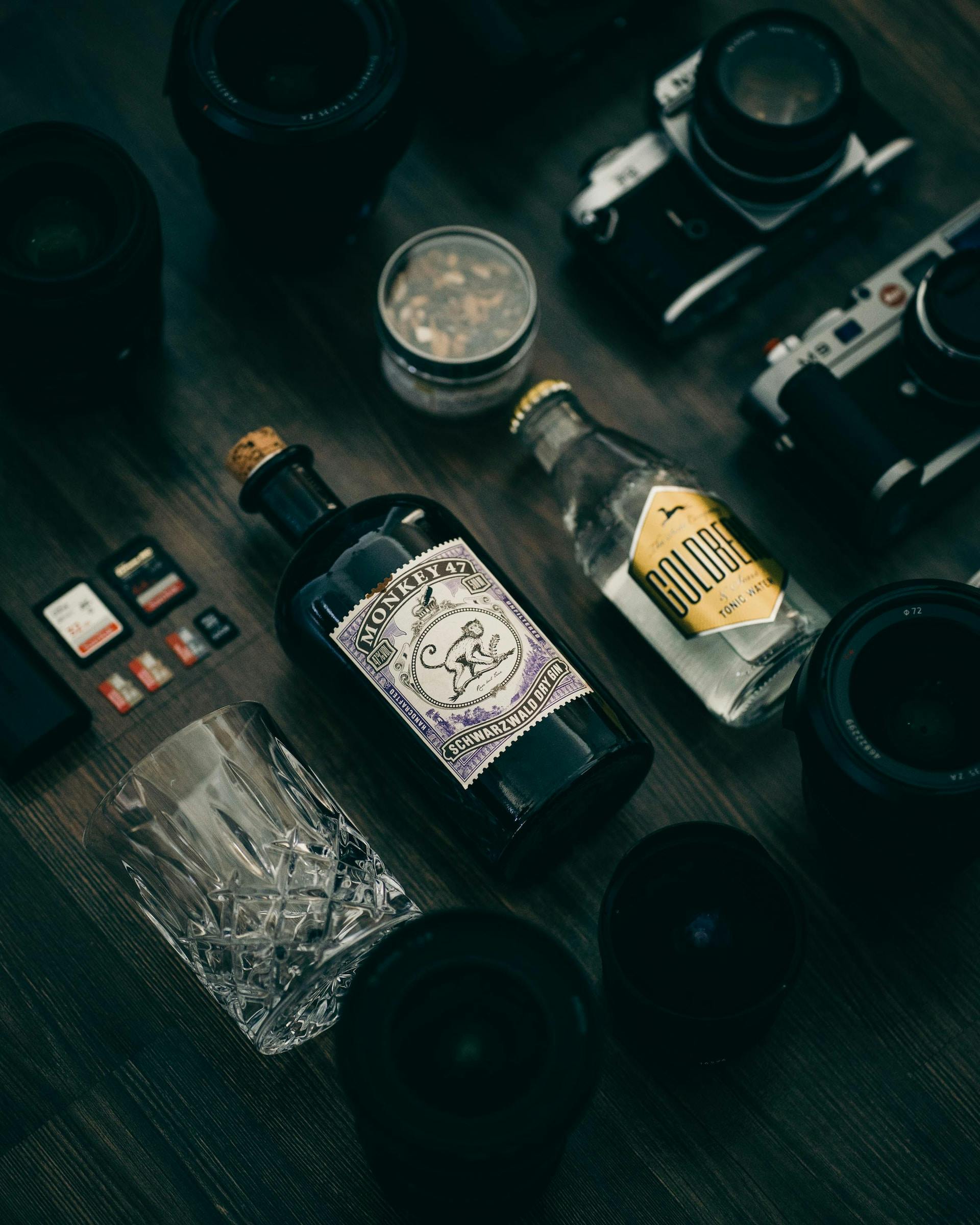 Auf dunklem Holzboden eine Flasche Gin, Tonic Water und ein Glas neben Kameras